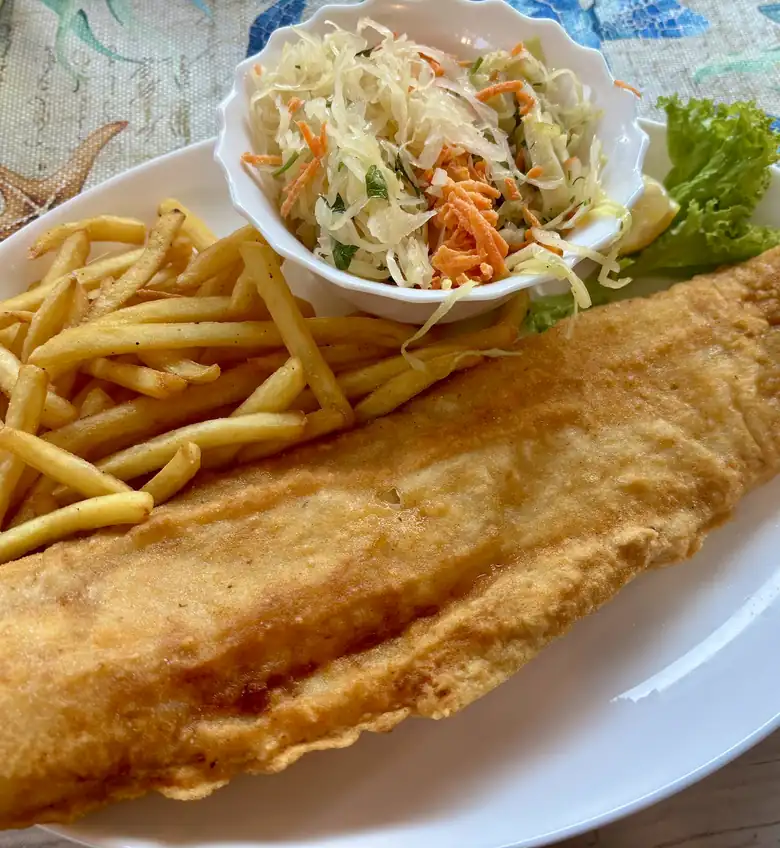 Duży filet ryby w złotej panierce z frytkami, surówką i sałatą na białym talerzu, na obrusie w niebieskie kwiaty